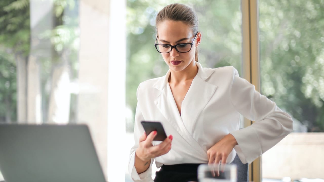 Lady in work clothes wearing glasses and blouse holding a phone in front of computer