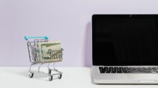 A small shopping cart full of cash sitting on a desk next to a MacBook